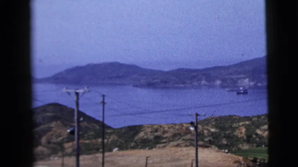 1958 8mm travel view across water toward low hills and distant mountains, with small boats and utility poles in the foreground.