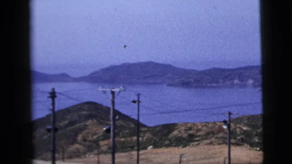 1958 8mm view of calm water, distant hills, and foreground power lines from a framed roadside or window perspective.