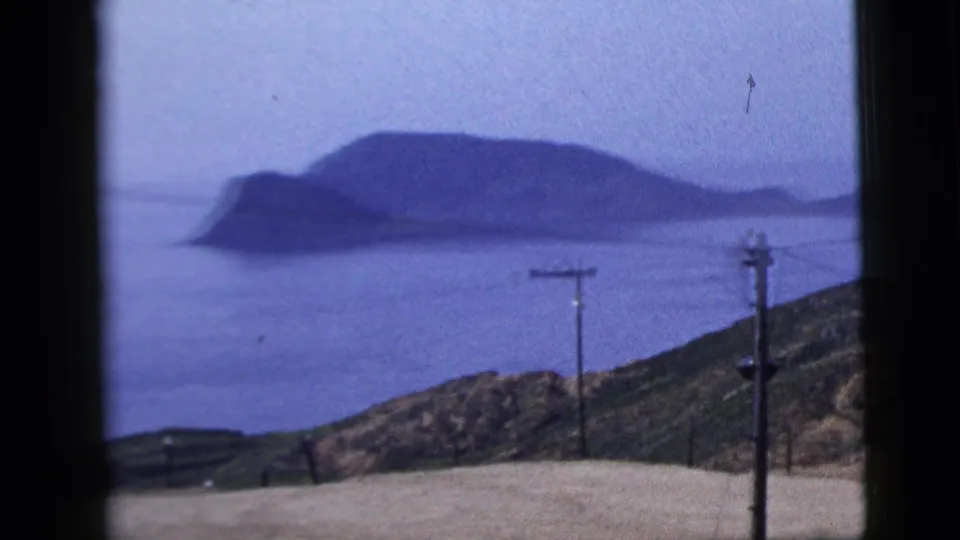 1958 8mm frame showing a hazy coastal headland across calm water, with roadside poles and sloping scrubland in front.