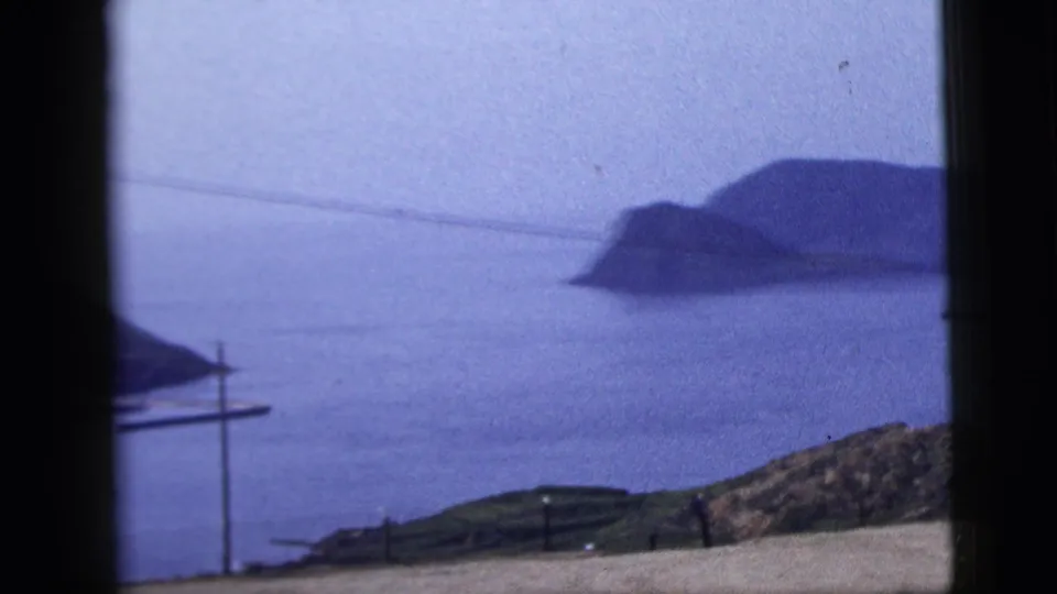 1958 8mm coastal view of a distant island or headland, calm water, foreground hills, and thin power lines.