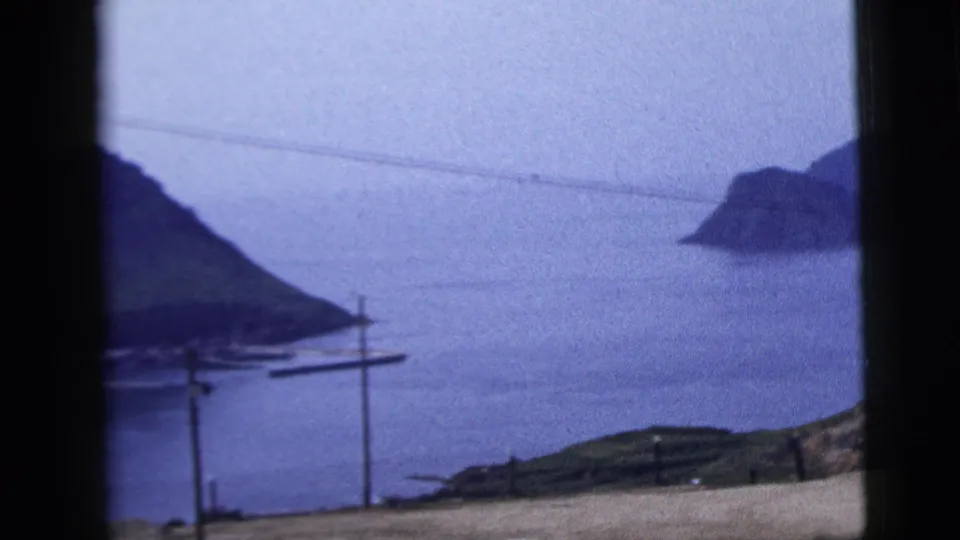 1958 8mm home-movie view of a calm harbor, distant landforms and utility poles framed by a dark vehicle or window edge.