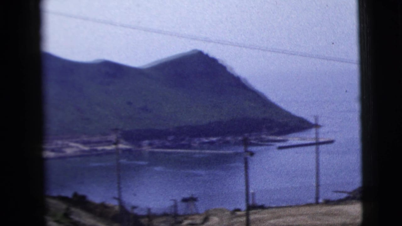 A coastal landform across calm water appears beyond foreground utility poles in this 1958 home-movie travel shot.