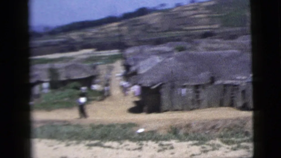 A small village with low buildings, a dirt road and a distant pedestrian appears below hills in the 1958 8mm footage.
