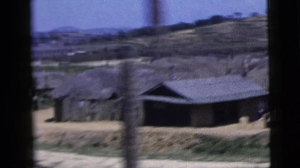 A moving 8mm view shows low rural buildings, a dirt path and hillside terrain, with people only indistinct if present.