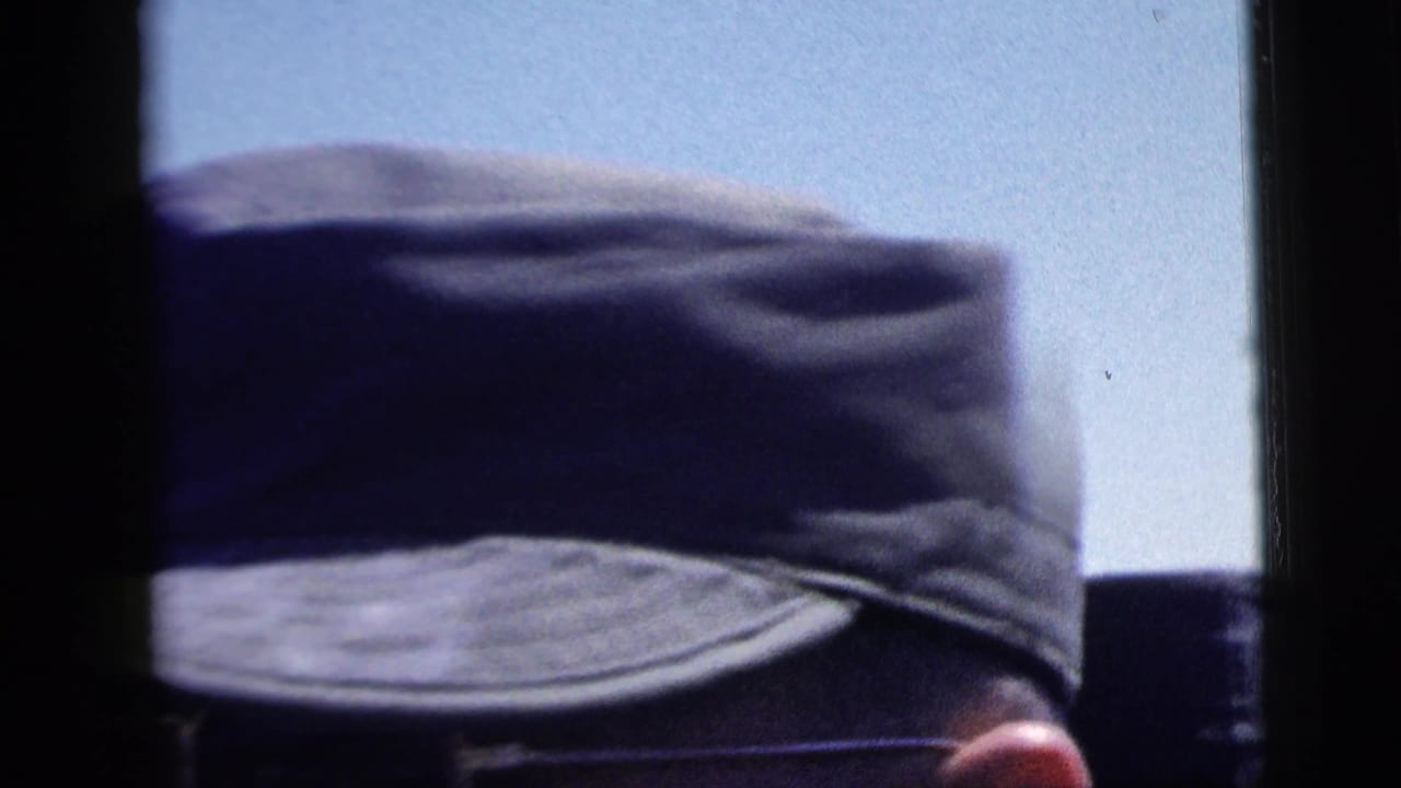 1958 New York City home-movie frame with a uniform cap in close foreground and a bright dockside background partly obscured.