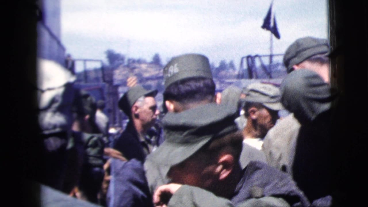 1958 New York City harbor footage shows uniformed men crowded on a ship deck, viewed through dark interior framing with a flag and shoreline beyond.