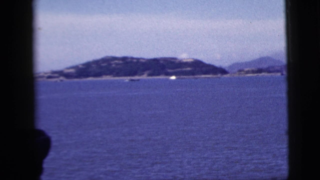 1958 8mm home-movie view from a vessel window toward blue water, hazy low shoreline, and small distant boats near New York City.
