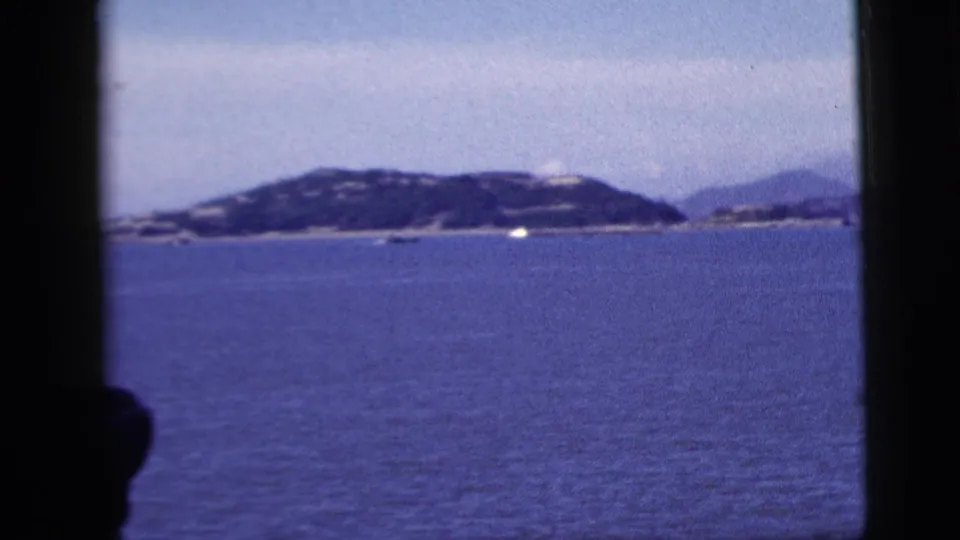 1958 8mm home-movie view from a vessel window toward blue water, hazy low shoreline, and small distant boats near New York City.