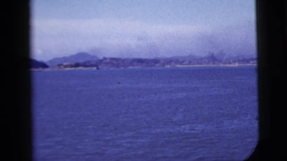 A soft 8mm frame shows blue water, a hazy distant coastline, and dark vessel-window edges during a 1958 coastal passage near New York City.