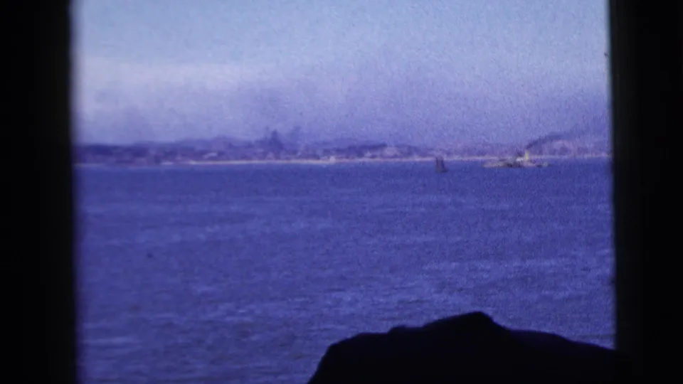Through the vessel window, blue water, small boats, and a distant hazy shoreline are visible, with a dark foreground shape entering the 8mm frame.