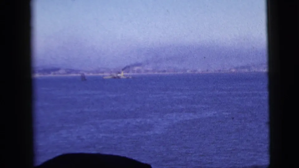 1958 New York City-area coastal water view with faint shoreline, possible small vessels, and dark window framing from onboard travel footage.