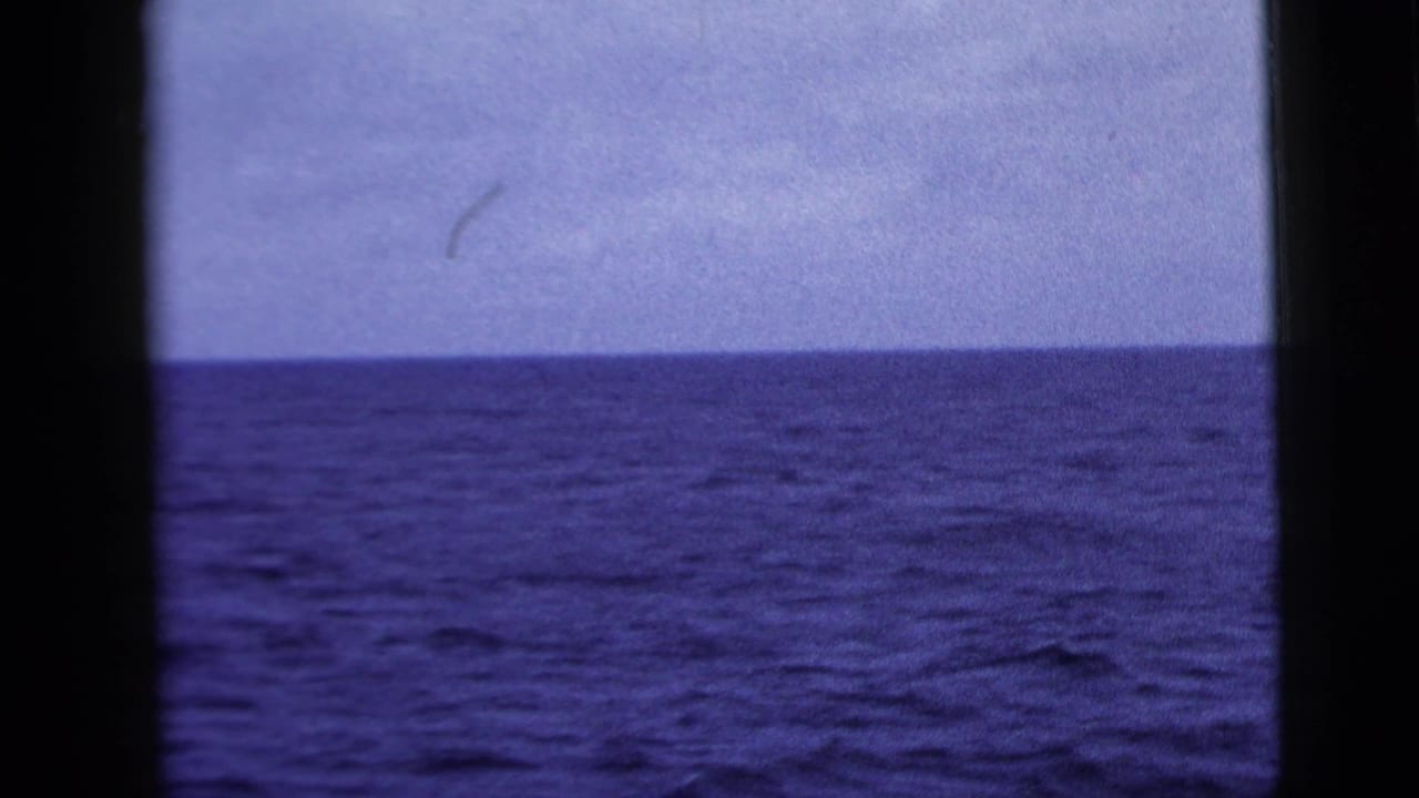 1958 8mm home-movie frame showing an empty blue ocean horizon through dark window-like bars, with cloudy sky above.