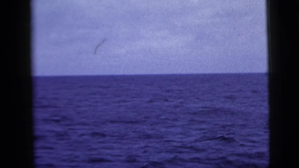 1958 8mm home-movie frame showing an empty blue ocean horizon through dark window-like bars, with cloudy sky above.