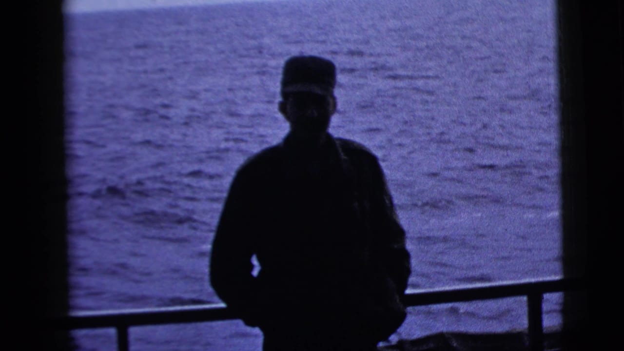 Silhouetted man in a cap stands on a boat deck with water behind him.