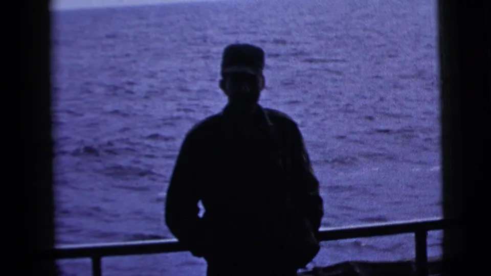 Silhouetted adult man in a military-style cap stands by a railing on a boat deck, with open water behind him in 1958 footage.