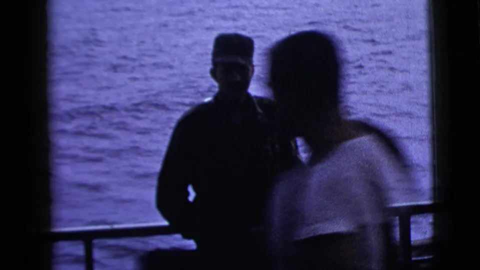 A silhouetted uniformed man stands near a boat railing while a blurred adult passerby crosses the foreground in 1958 8mm footage.