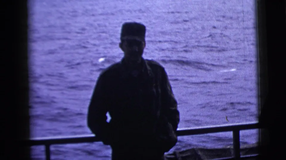 1958 8mm frame of a silhouetted adult man in a cap standing by a boat railing with rippled water in the background.