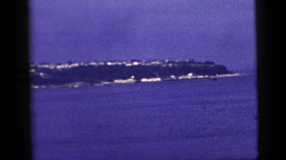 A distant shoreline and low cluster of buildings appear across the water, seen from a boat in the 1958 New York City home-movie clip.