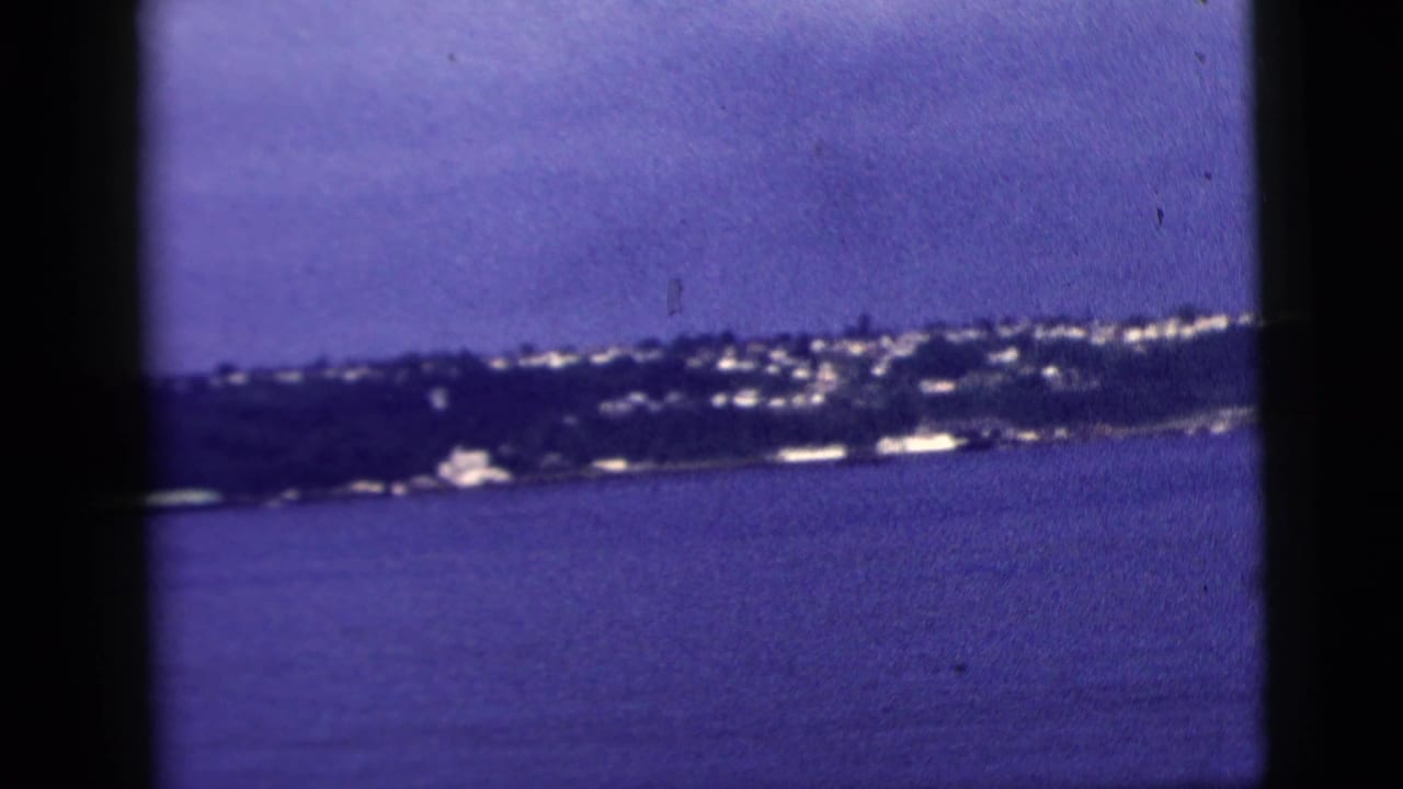 1958 New York City home-movie frame showing a distant shoreline across water, softened by blue-purple film color shift.