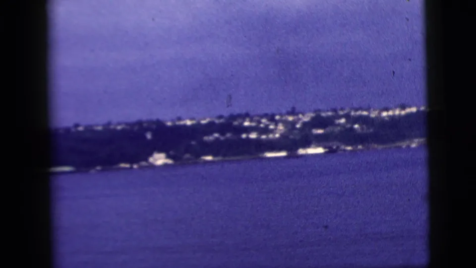 1958 New York City home-movie frame showing a distant shoreline across water, softened by blue-purple film color shift.