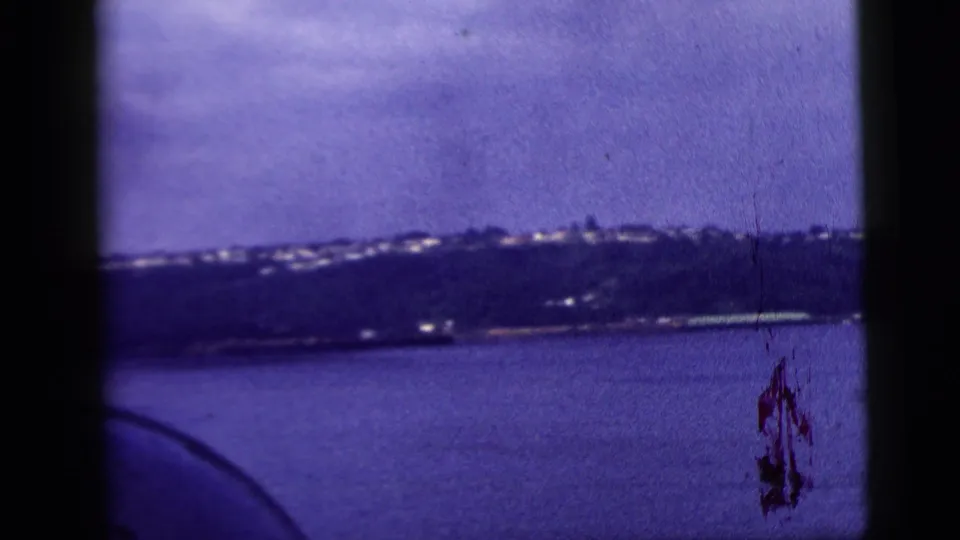 Home-movie view across water toward a distant shore, with a dark curved foreground form entering the frame.