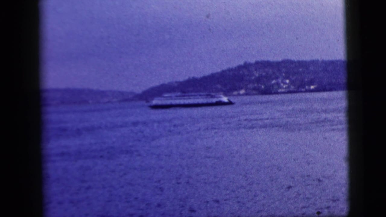 A large ferry-like vessel crosses New York City waters in a 1958 8mm frame, seen past dark window dividers and a populated hillside.