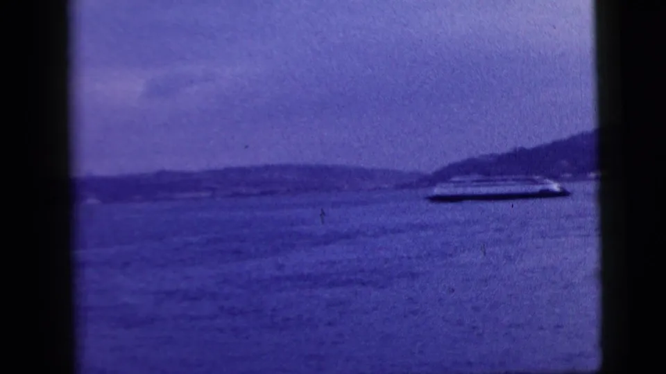 The 1958 home-movie frame shows a ferry-like boat on New York City waters, with distant shoreline and dark window edges in the foreground.