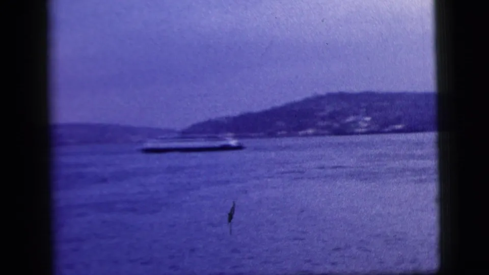 1958 8mm New York City harbor footage with a small boat on the water and a distant hilly shoreline beyond it.