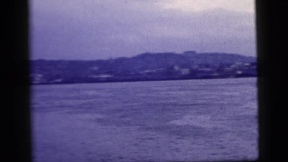 Wide 1958 New York City harbor view from a boat, with water in the foreground and a hazy distant shoreline across the frame.