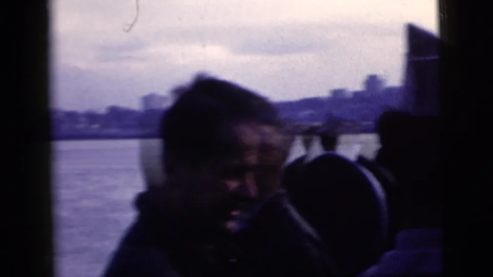 Silhouetted passengers aboard a boat in 1958 New York City harbor footage, including a man in a military-style cap facing the shoreline.
