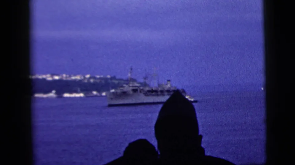 1958 New York City 8mm harbor frame showing a vessel on blue water, foreground silhouettes, and a lit distant shoreline.