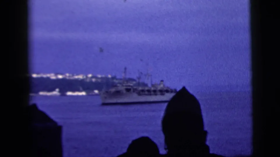Blue-toned 8mm frame of a large ship offshore in New York City, with dark human silhouettes watching from the foreground.