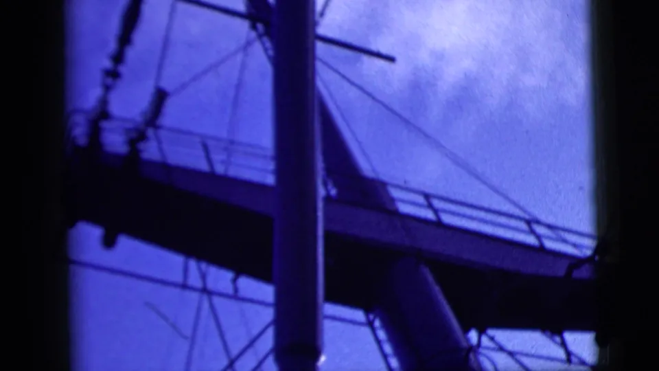 Close-up 8mm view of a ship mast, rigging, and deck structure against a blue, clouded sky.