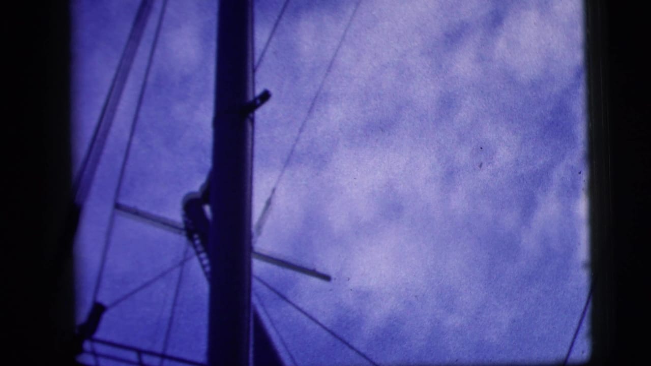 Blue-tinted 8mm frame with dark borders, showing sky and angled structural or window lines before the outdoor uniformed-group scene.