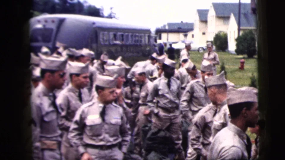 1958 New York City home-movie frame showing uniformed men in grey outfits and pale caps gathered near a bus and white clapboard buildings.