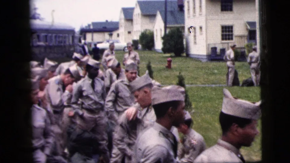 1958 New York City 8mm frame with uniformed men on a grassy lawn, a bus at left, white buildings, and a red fire hydrant in the background.