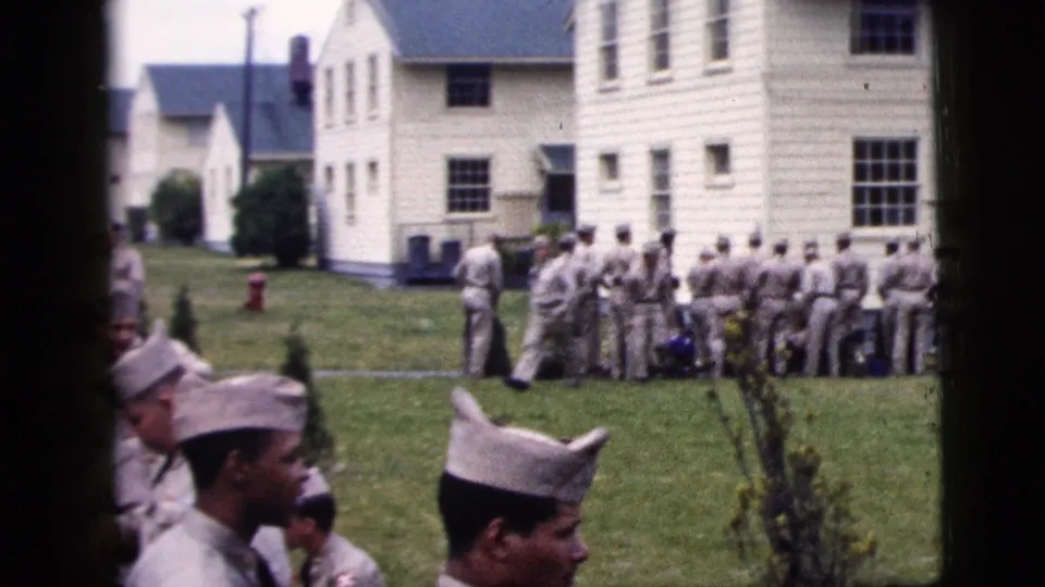 1958 New York City home-movie frame showing uniformed men lined up near white clapboard buildings, with several figures closer to the camera.
