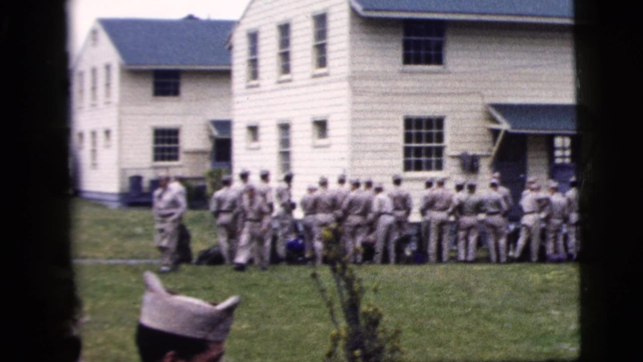 1958 home-movie frame of men in grey uniforms standing on a lawn outside a white clapboard building in New York City metadata context.