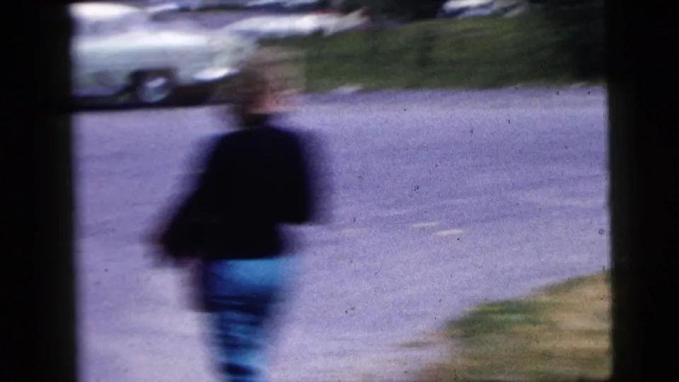 Handheld 1958 home-movie view of a blurred pedestrian passing a roadway with a parked car in the background.