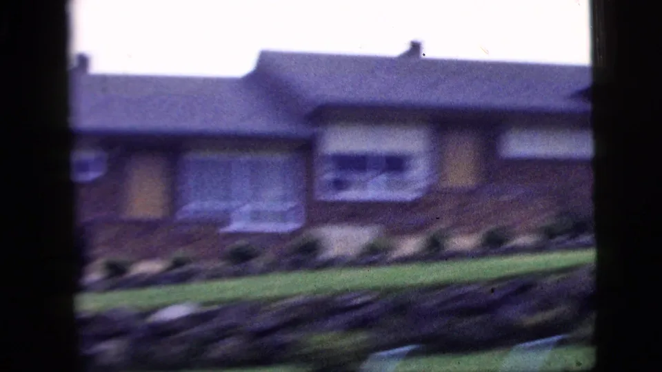 Soft-focus 1958 home-movie frame showing a low brick residential house with windows, shrubs, and a front lawn.