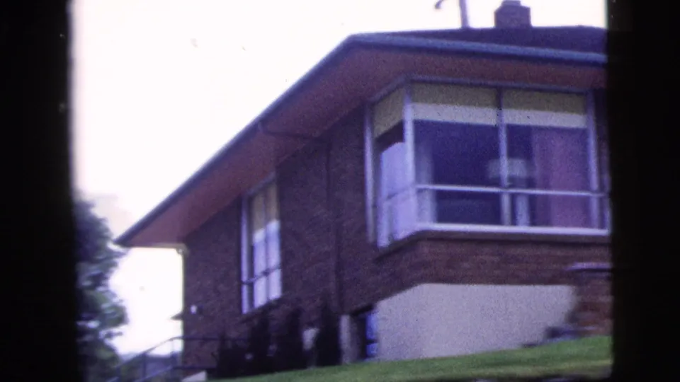 1958 home-movie frame of a brick residential exterior with broad corner windows and a sloping lawn.