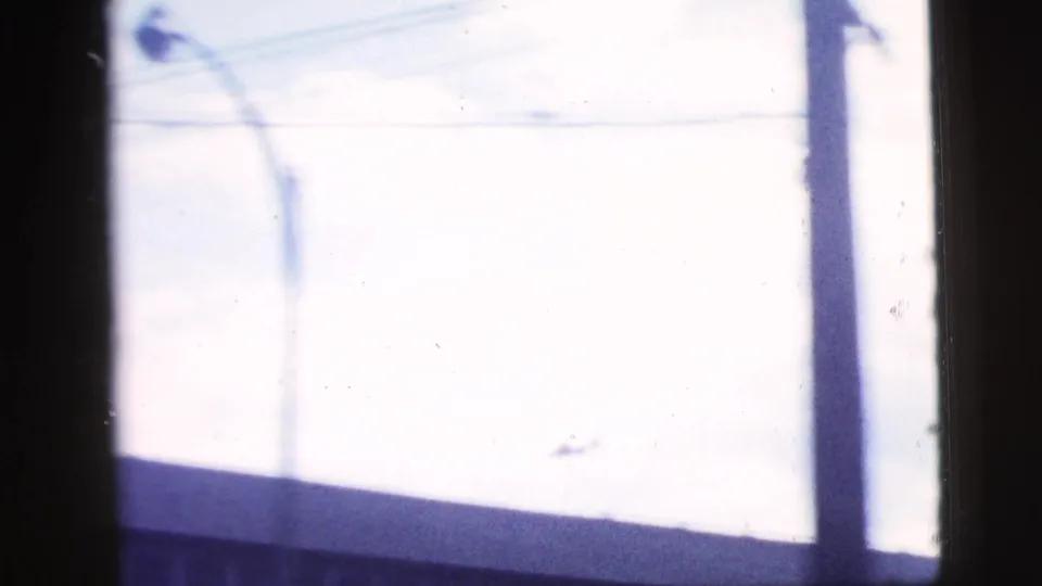 A small airplane appears in the bright sky above a New York City roofline and utility pole in 1958 8mm footage.