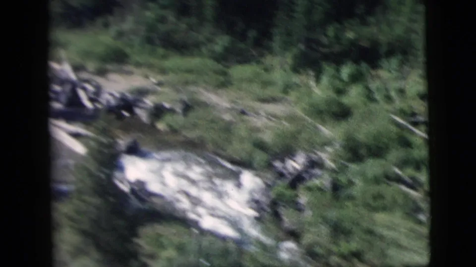 Moving-train 8mm frame showing a rocky stream bed, pale rushing water, and surrounding green vegetation.