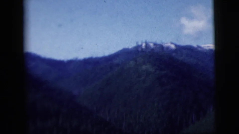 1958 8mm frame of a dark forested mountain ridge with small snow or light patches under blue sky.