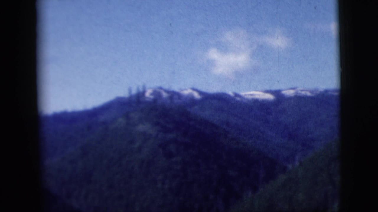 1958 8mm home-movie frame showing mountains and evergreen slopes through a dark vehicle window divider.
