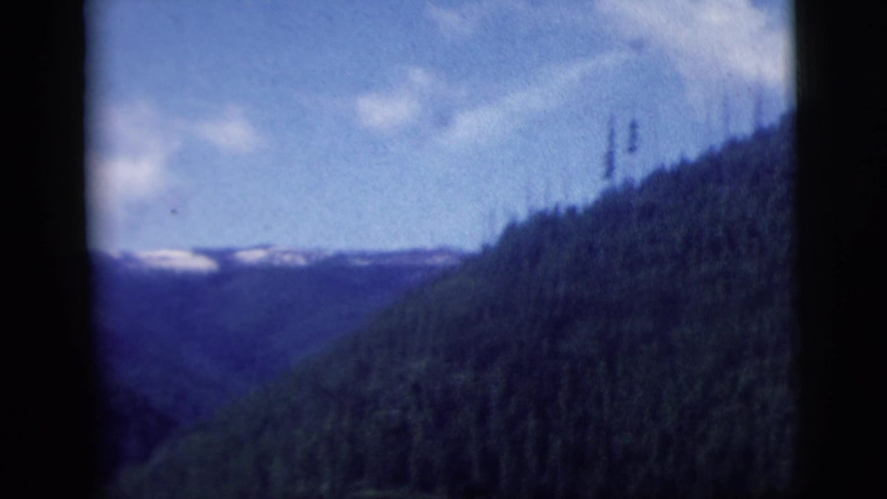 Passenger-window view of a mountain valley, evergreen hillside, and distant snow patches in 1958 home-movie footage.