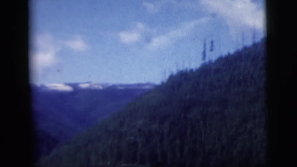 Passenger-window view of a mountain valley, evergreen hillside, and distant snow patches in 1958 home-movie footage.
