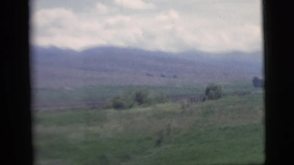 1958 8mm travel frame showing open fields, scattered trees, and hazy hills through a vehicle window.