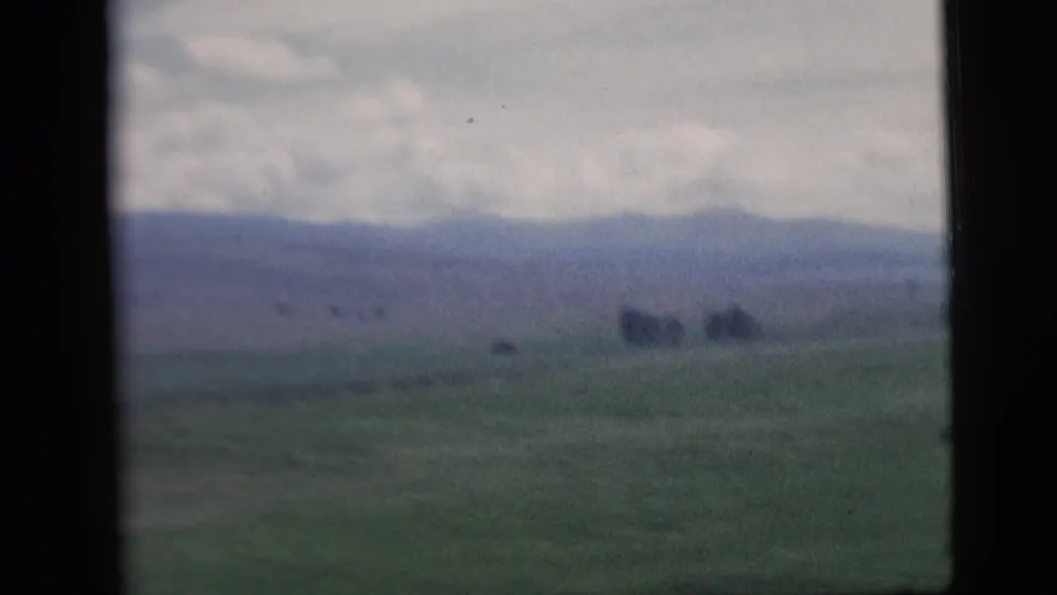 Passenger view across farmland toward distant hills, framed by a vehicle window in faded 1958 8mm footage.