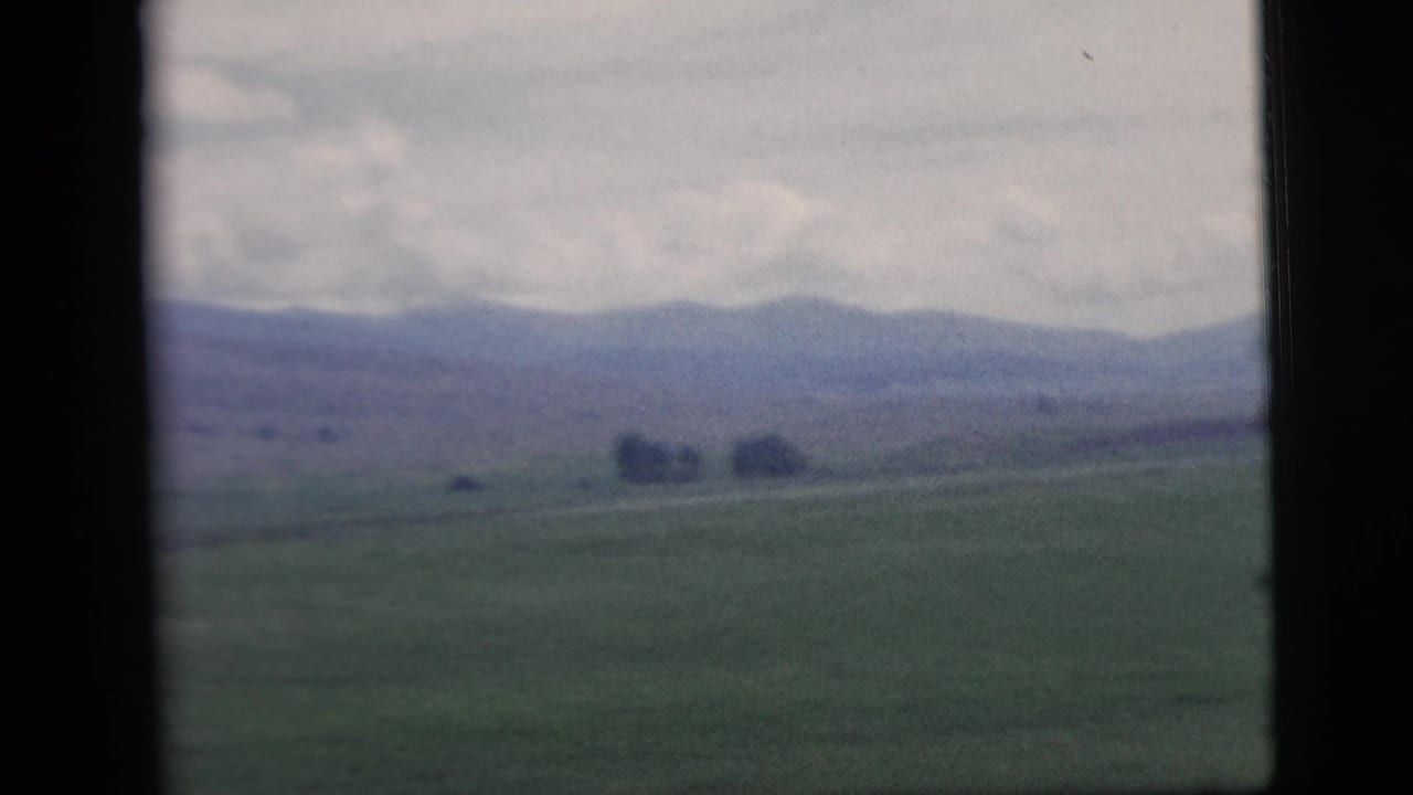 1958 home-movie view of rural fields and distant hills seen through a window, with soft focus and visible 8mm frame edges.